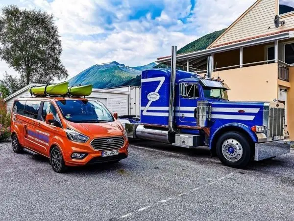 Zwei Fahrzeuge stehen auf einem Parkplatz, ein orangefarbener Van mit Kanus und ein blauer Lkw