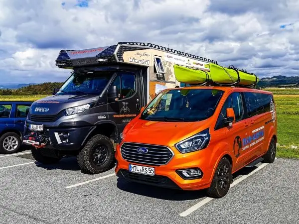 Zwei Fahrzeuge stehen auf einem Parkplatz, ein schwarzer Offroad-Wohnmobil und ein orangefarbener Van mit einem Kajak auf
