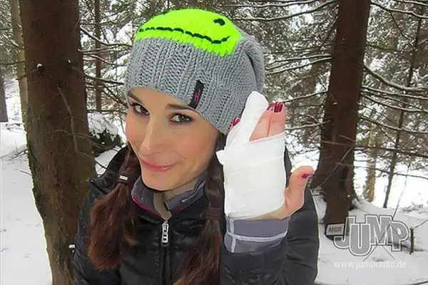 Eine Person mit einer grauen Mütze und verbundenem Handgelenk lächelt in einem Wald