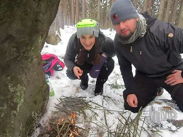 Kleine Flamme brennt zwischen Ästen im Schnee, zwei Personen knien daneben