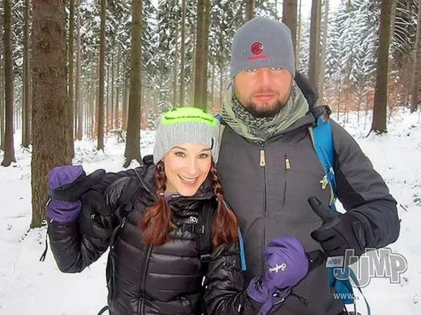 Zwei Personen posieren fröhlich im verschneiten Wald, umgeben von hohen Bäumen
