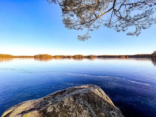 Großer Stein am Ufer eines ruhigen Gewässers mit Bäumen im Hintergrund
