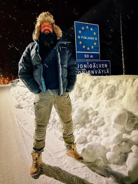 Mann in warmer Jacke und Stiefeln steht vor einem Schild mit der Aufschrift „Finland“