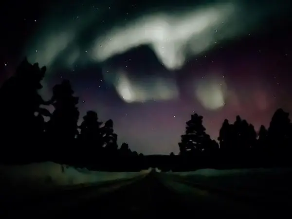 Sternenhimmel mit bunten Lichtspielen über einer schneebedeckten Straße und silhouettierten Bäumen