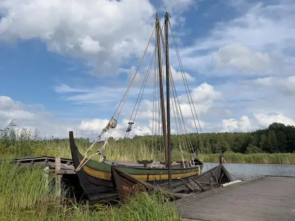 Holzboot mit Segeln liegt an einem Steg, umgeben von Schilf und Wasser