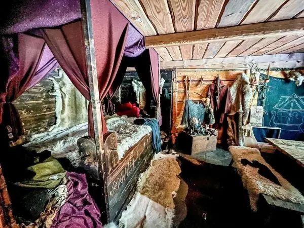 Innenansicht eines rustikalen Raumes mit einem Bett, Fellen und Kleidung