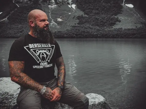 Mann mit tätowierten Armen sitzt am Ufer eines Sees vor einer Berglandschaft