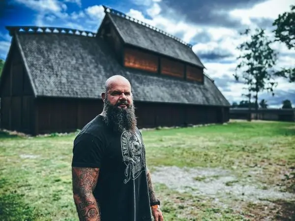 Mann mit tätowierten Armen steht vor einem traditionellen Holzgebäude