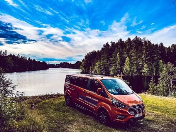 Oranger Van steht am Ufer eines ruhigen Gewässers, umgeben von Bäumen