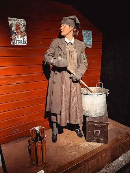 Person in historischer Kleidung rührt in einem Topf, Holzwand im Hintergrund