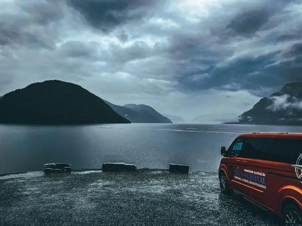 Roter Van am Ufer eines ruhigen Sees mit Bergen im Hintergrund