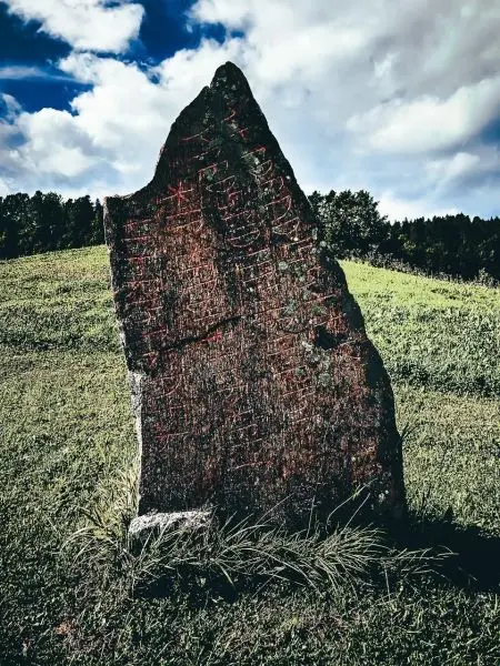 Steinerne Stele mit eingravierten Mustern auf einer Wiese unter bewölktem Himmel
