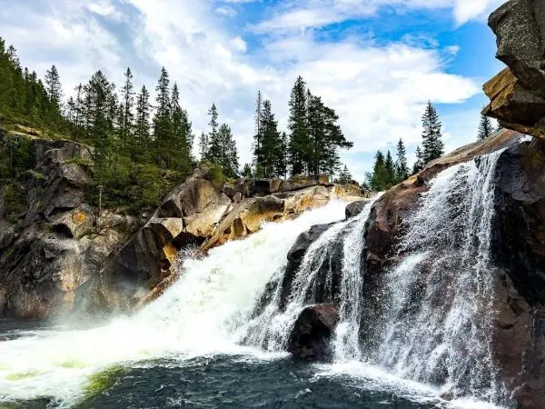 Wasserfall stürzt über Felsen in einen klaren See, umgeben von Bäumen