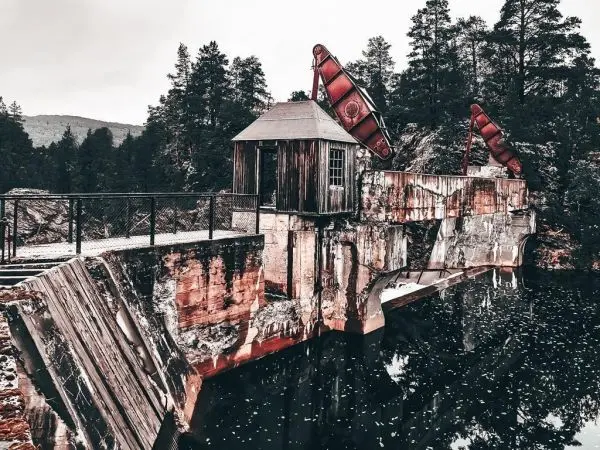 Alter Staudamm mit Holzgebäude und roten Klappen, umgeben von Bäumen
