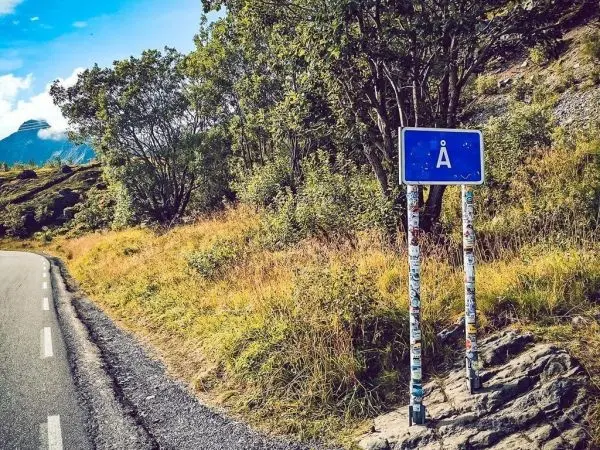Blaues Ortsschild mit dem Buchstaben „Å“ an einer Straße, umgeben von Bäumen und Gras