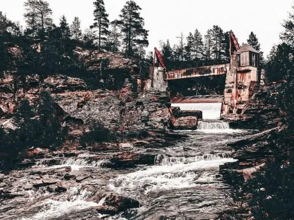 Blick auf eine alte Staumauer mit Wasserfall und umgebenden Bäumen