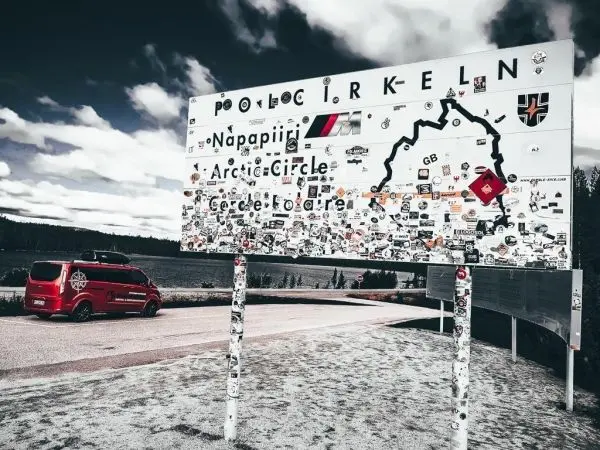 Hinweisschild mit Aufschriften und Aufklebern, rotes Fahrzeug parkt daneben