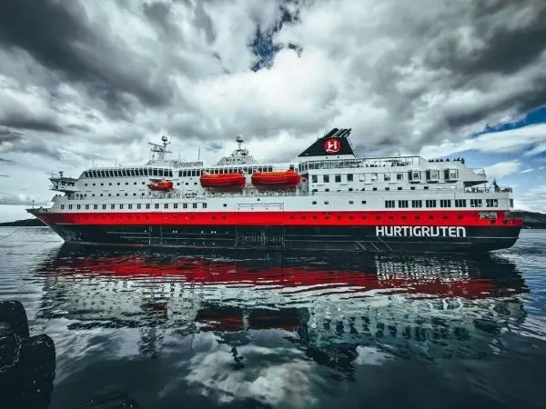 Kreuzfahrtschiff mit rotem und weißem Rumpf spiegelt sich im Wasser unter bewölktem Himmel