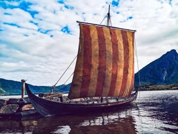 Langschiff mit gestreifter Segel steht am Ufer eines Gewässers