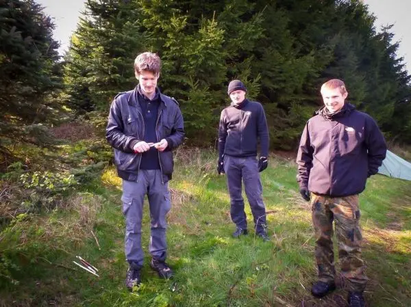 Drei Personen stehen im Freien und betrachten einen Stock auf dem Boden