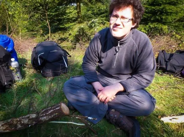 Mann sitzt im Gras neben einem Holzstück und Rucksack