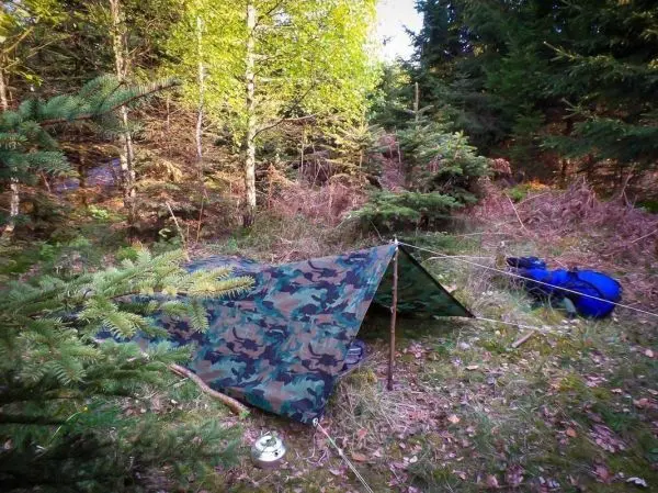Tarp-Notunterkunft aus Tarnmaterial zwischen Bäumen, Rucksack daneben