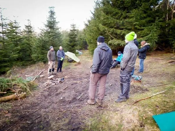 Gruppe steht um ein kleines Bodenfeuer im Wald, Tarp-Notunterkunft im Hintergrund
