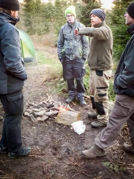 Kleine Flamme brennt auf einem Holzstamm, während Personen um das Feuer stehen
