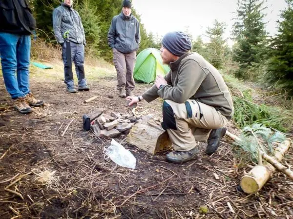 Mann erklärt Techniken am Lagerfeuer, Holz und Ausrüstung liegen auf dem Boden