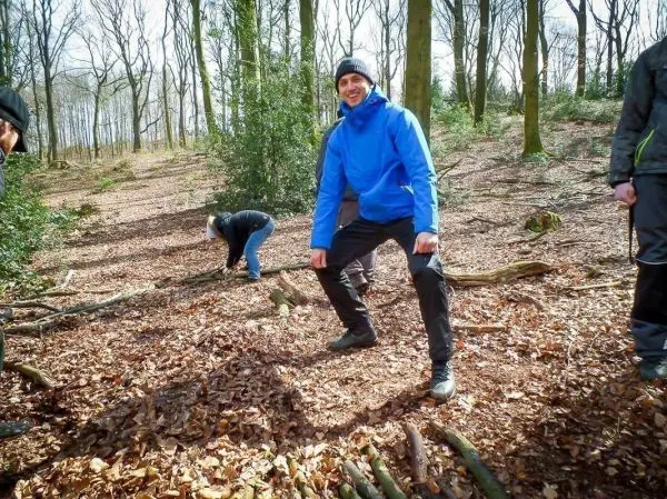 Mann in blauer Jacke steht auf Laub, während andere Holzstücke sammeln