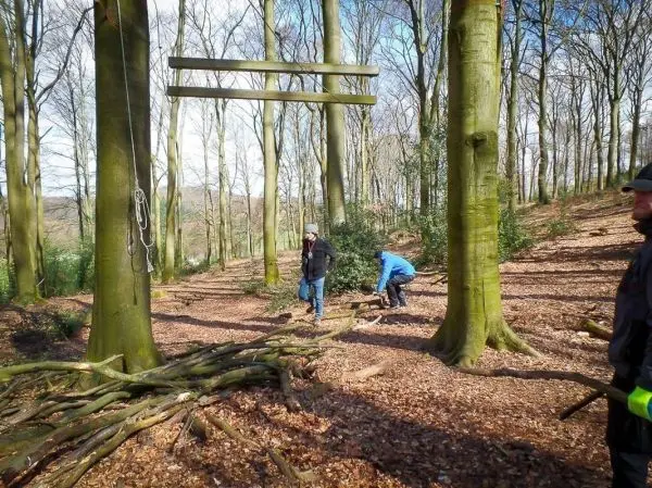 Personen stehen unter Bäumen neben einer selbstgebauten Seilbrücke