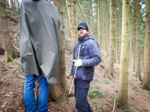 Zwei Personen stehen im Wald, einer hält einen Stock und schaut zur anderen