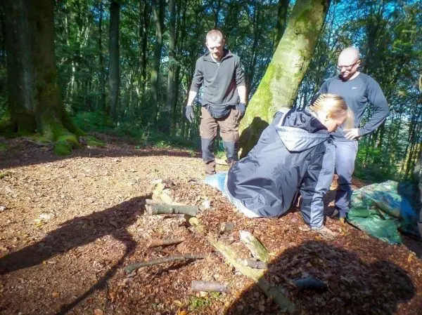 Drei Personen arbeiten im Wald an einer improvisierten Tragekonstruktion aus Ästen