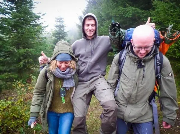 Drei Personen tragen Ausrüstung und gehen durch einen bewaldeten Bereich