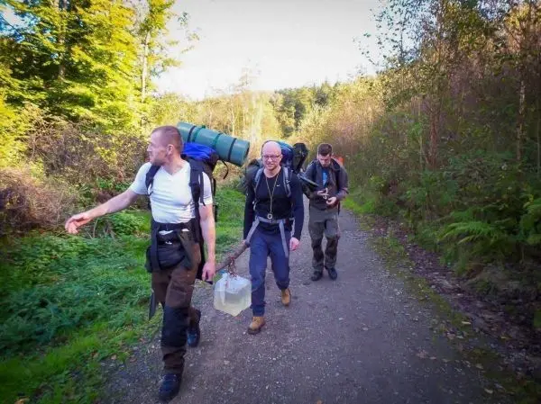 Drei Personen wandern mit Rucksäcken auf einem Waldweg beim Überlebenstraining