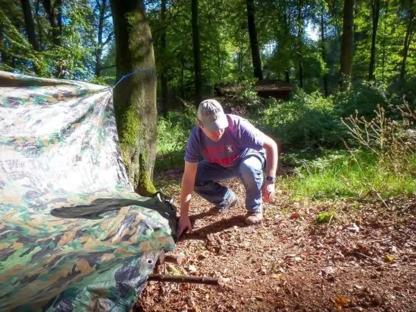 Mann richtet Tarp-Notunterkunft mit Holzstück und Seil aus