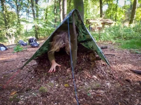 Person kriecht aus einem selbstgebauten Unterschlupf im Wald