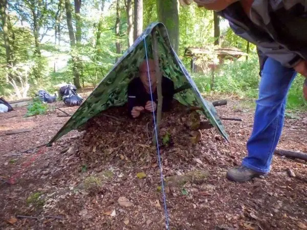 Person sitzt unter einer selbstgebauten Plane im Wald
