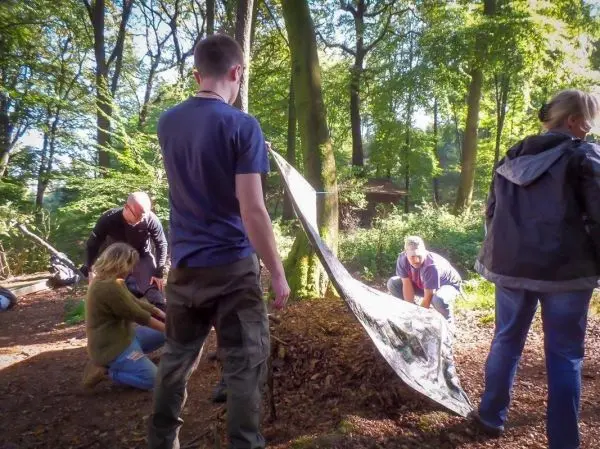 Tarp-Notunterkunft wird zwischen Bäumen gespannt, Teilnehmer arbeiten zusammen am Aufbau