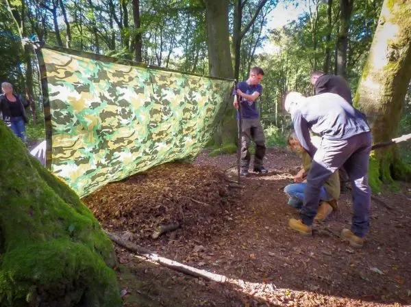 Tarp-Notunterkunft wird zwischen Bäumen gespannt, während Teilnehmer den Untergrund vorbereiten