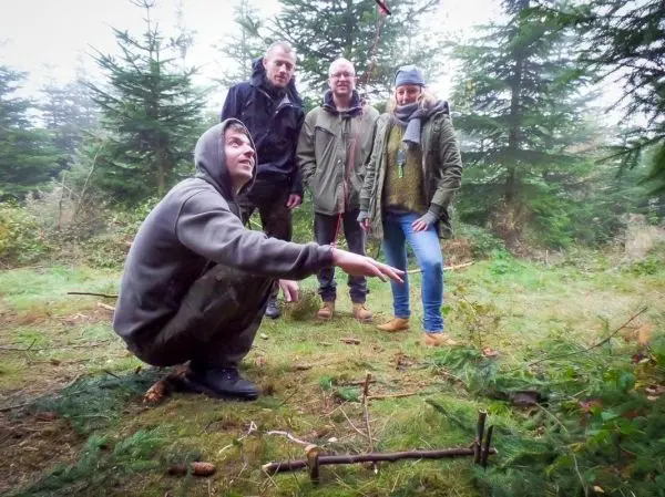 Vier Personen stehen in einem Wald, während eine Person auf dem Boden sitzt und auf einen Stock zeigt