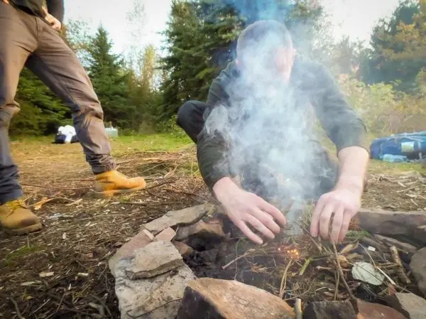 Zunder glimmt im Feuerkreis, Rauch steigt auf, Hände fügen Äste hinzu