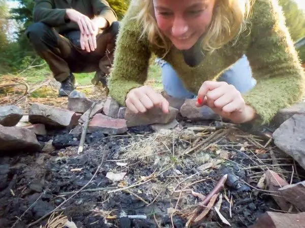 Zunder glimmt in einem Feuerkreis aus Steinen, Hände nähern sich vorsichtig