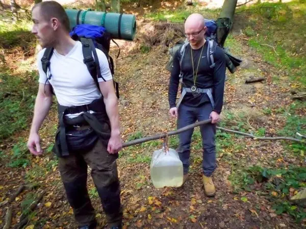 Zwei Personen tragen einen Stock mit einem Wasserbehälter durch den Wald