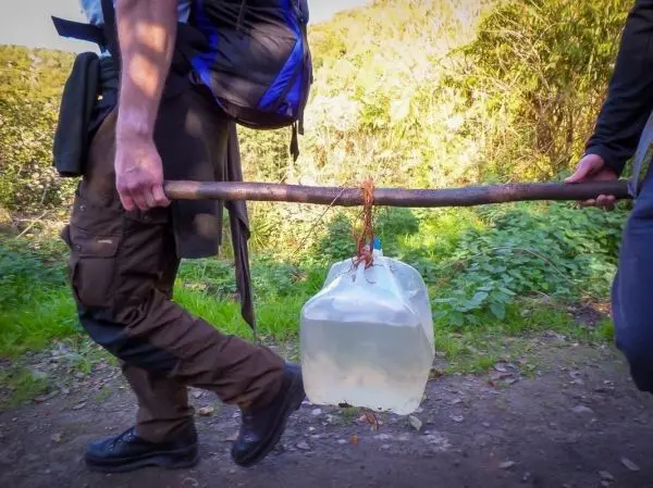 Zwei Personen tragen einen Wasserbehälter an einem Stock entlang eines Weges