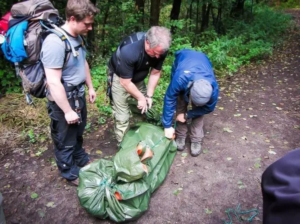 Drei Personen sichern eine grüne Tragetasche mit einem Seil im Wald
