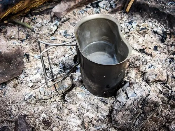 Edelstahlkessel steht auf grauen Asche-Resten eines Feuers