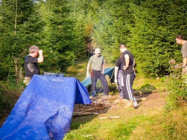 Gruppe steht um ein kleines Bodenfeuer, Tarp-Notunterkünfte im Hintergrund