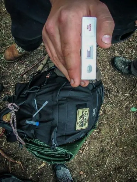 Hand hält ein Testgerät über einem Rucksack mit Ausrüstung und Seil