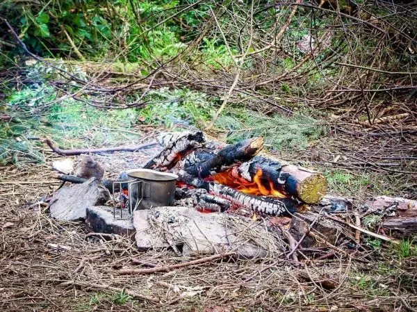 Kleine Flamme brennt zwischen Holzscheiten, Kessel steht auf Steinen